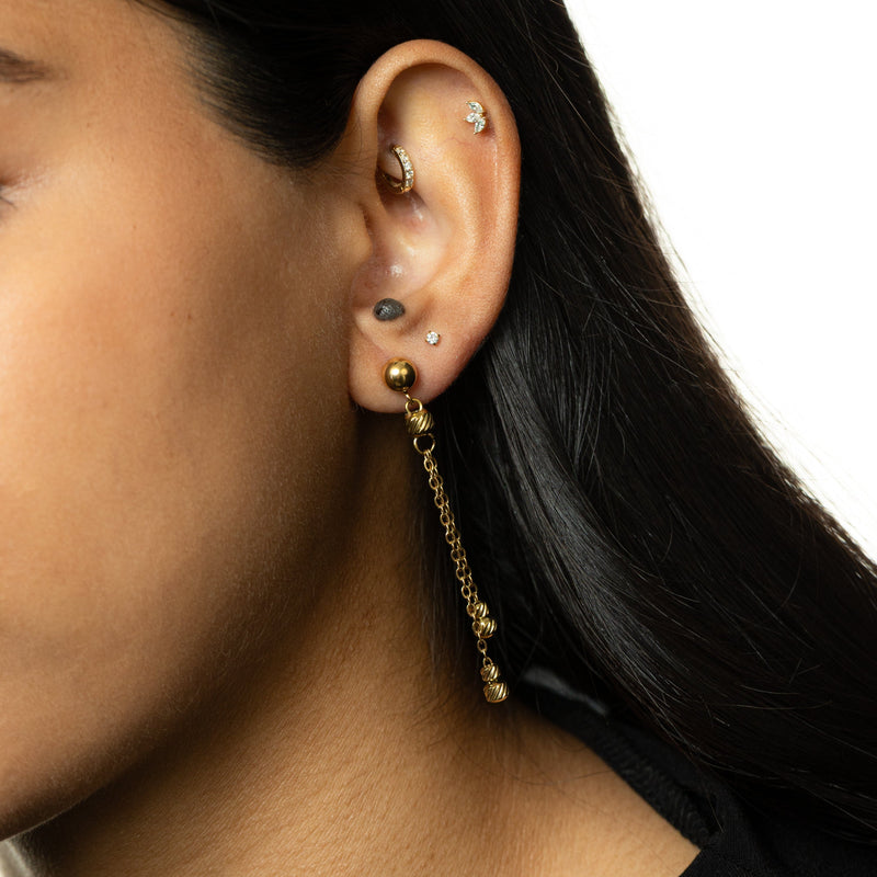 Noor Chain Drop Earrings — Woman wearing a single gold earring with double chain dangles ending in ribbed bead charms, styled with black hair tucked back.